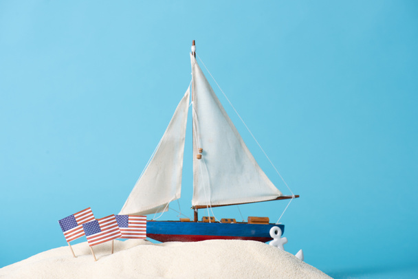 decoratieve schip in de buurt van Amerikaanse nationale vlaggen in wit zand geïsoleerd op blauw - Foto, afbeelding