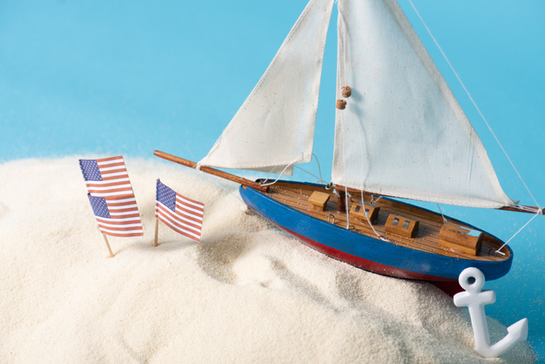 Banderas nacionales americanas en arena blanca cerca de barco en miniatura y ancla aislada en azul
 - Foto, Imagen
