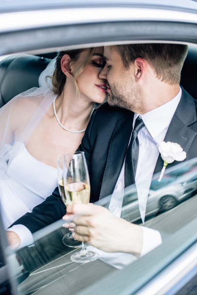 atraktivní nevěsta a pohledný ženich líbání a držení skleničky šampaňského - Fotografie, Obrázek