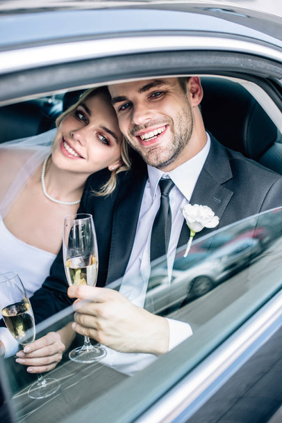 ελκυστική νύφη και όμορφος γαμπρός χαμογελώντας και κρατώντας γυαλιά σαμπάνιας - Φωτογραφία, εικόνα