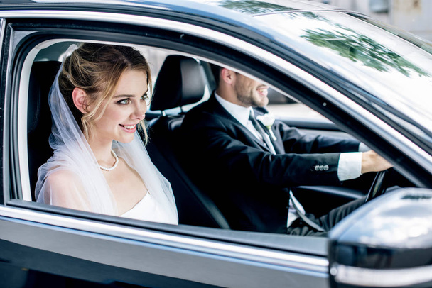 vonzó menyasszony menyasszonyi fátyol és a vőlegény mosolyogva az autóban  - Fotó, kép