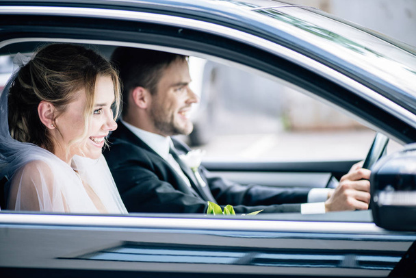 επιλεκτική εστίαση της ελκυστική νύφη σε νυφικό πέπλο και γαμπρός χαμογελά στο αυτοκίνητο  - Φωτογραφία, εικόνα