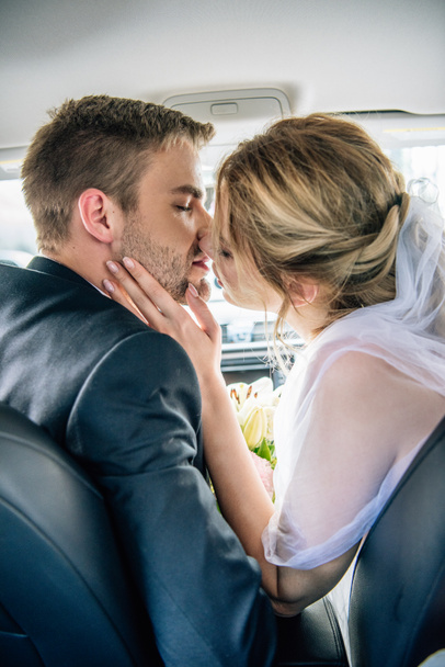 πίσω όψη της ελκυστική νύφη σε νυφικό πέπλο και γαμπρός φιλιά στο αυτοκίνητο  - Φωτογραφία, εικόνα