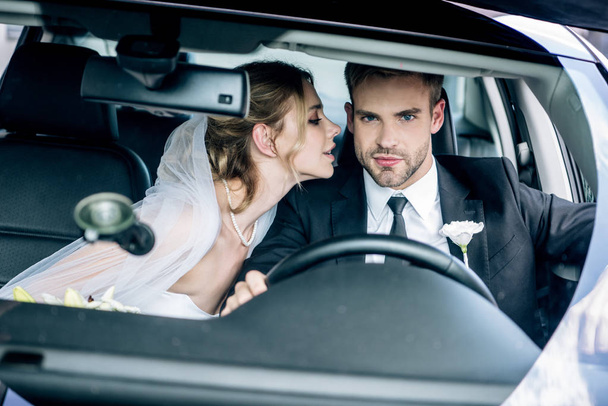 attractive bride in bridal veil kissing handsome bridegroom in car  - Photo, Image