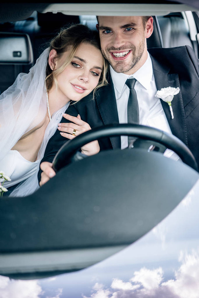 ελκυστική νύφη σε νυφικό πέπλο αγκαλιάζει όμορφος γαμπρός στο αυτοκίνητο  - Φωτογραφία, εικόνα