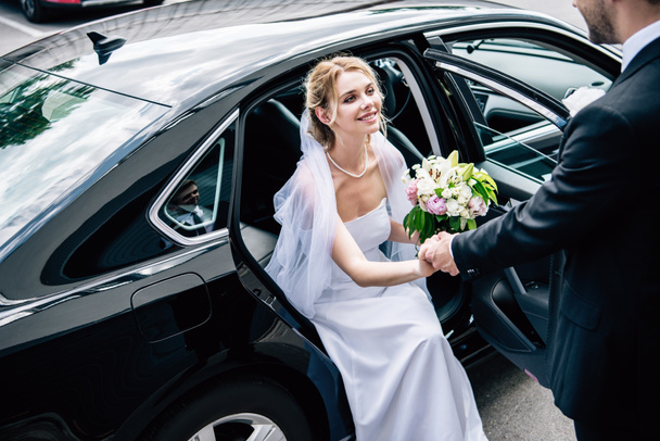 γαμπρός σε κοστούμι δίνοντας το χέρι σε ελκυστική και χαμογελαστή νύφη με ανθοδέσμη - Φωτογραφία, εικόνα