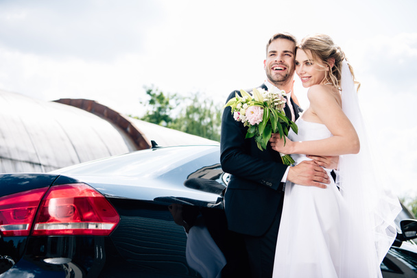 handsome bridegroom in suit hugging attractive and blonde bride with bouquet - Fotografie, Obrázek