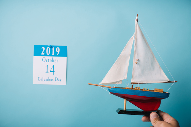 περικομμένη όψη του ανθρώπου που κατέχει κατασκευασμένο πλοίο κοντά σε χαρτί ημερολόγιο με επιγραφή ημέρα Κολόμπους απομονωμένη σε μπλε - Φωτογραφία, εικόνα