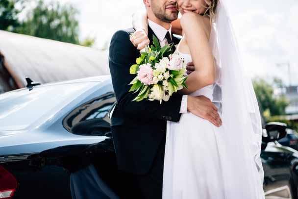 περικομμένη όψη του γαμπρού στο κοστούμι αγκάλιασμα νύφη σε νυφικό με ανθοδέσμη - Φωτογραφία, εικόνα