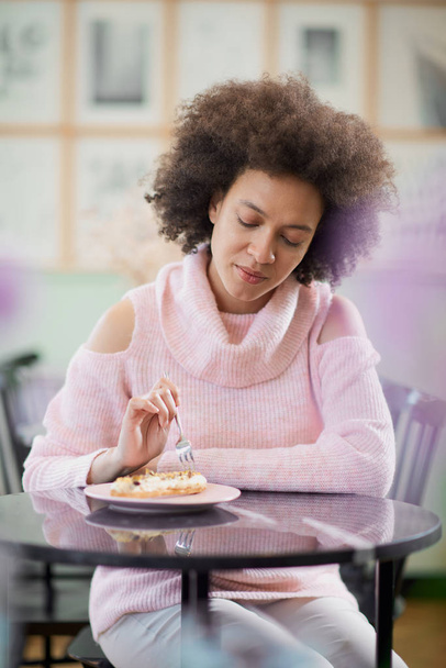Porträt einer charmanten positiven Mischlingsfrau im rosafarbenen Rollkragenpullover, die in einer Konditorei sitzt und Torte isst. - Foto, Bild