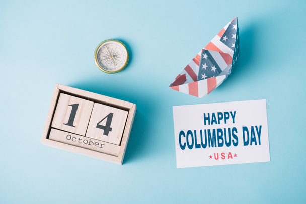 top view of calendar with October 14 date near paper boat with American flag pattern, compass and card with happy Columbus Day inscription on blue background  - Photo, Image