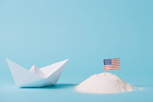 papier boot in de buurt van de Amerikaanse nationale vlag in wit zand op blauwe achtergrond - Foto, afbeelding