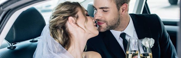 panoramic shot of attractive bride and handsome bridegroom kissing and holding champagne glasses - Foto, Bild
