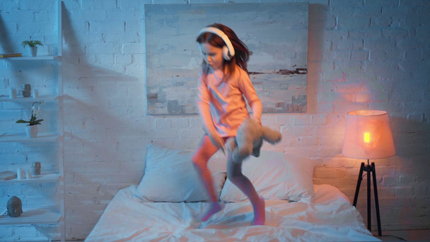 child in headphones jumping on bed at night - Filmati, video