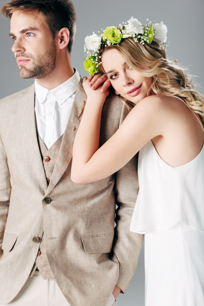 bride in wedding dress and wreath hugging handsome bridegroom in suit isolated on grey - Photo, Image