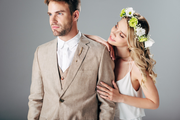 bride in wedding dress and wreath hugging handsome bridegroom in suit isolated on grey - Photo, Image