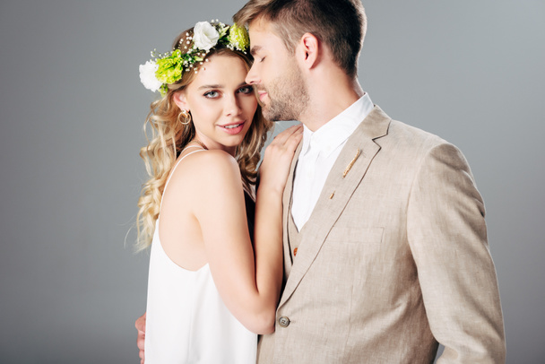handsome bridegroom in suit hugging with bride in wedding dress and wreath isolated on grey - Фото, зображення
