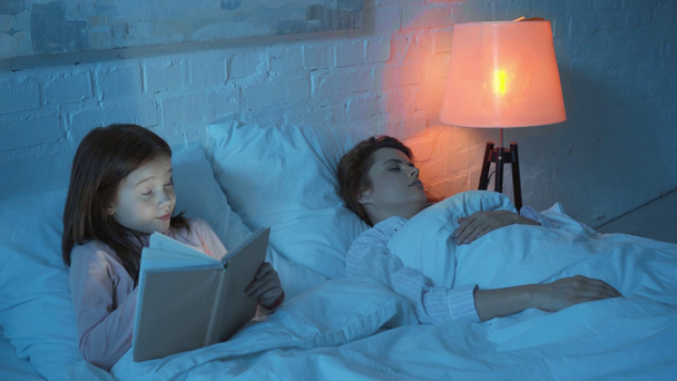 mother sleeping and daughter reading book with flashlight in bed - Imágenes, Vídeo