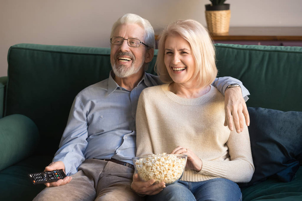Happy senior couple holding remote control snack laughing watching tv - Фото, изображение