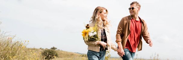 Panoramaaufnahme einer attraktiven Frau mit Blumenstrauß und einem hübschen Mann, der lächelt und Händchen hält  - Foto, Bild