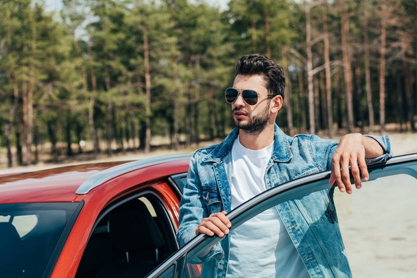handsome man in sunglasses and denim jacket standing near car - Foto, Imagem