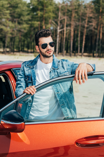 handsome man in sunglasses and denim jacket standing near car - Zdjęcie, obraz