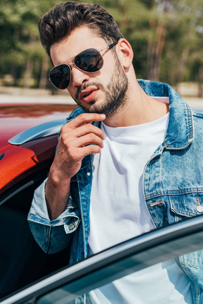 handsome man in sunglasses and denim jacket standing near car - Foto, afbeelding