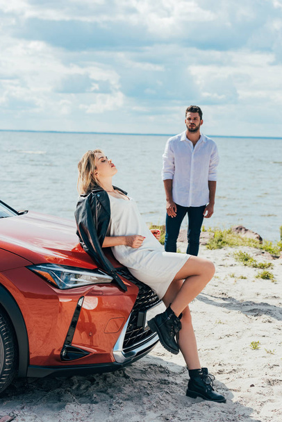 attractive woman in dress with closed eyes lying on car and man look at her  - Zdjęcie, obraz