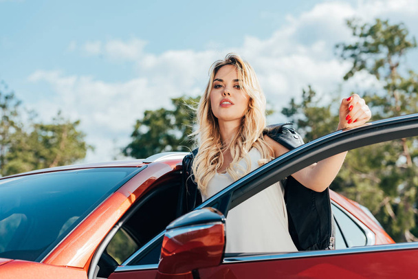 attractive and blonde woman standing near car and looking at camera  - Valokuva, kuva