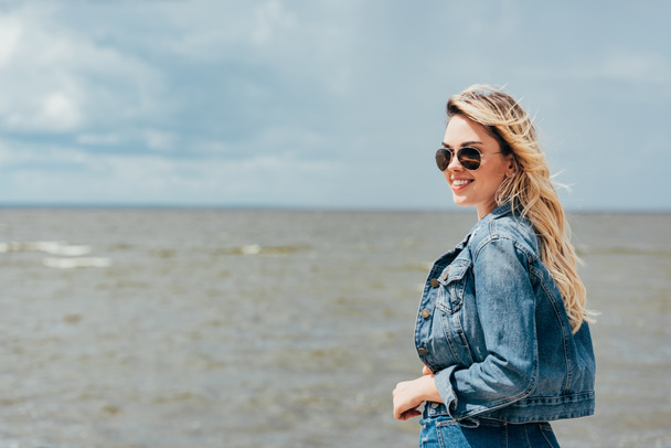 blonde and attractive woman in denim jacket and sunglasses looking away  - Фото, изображение