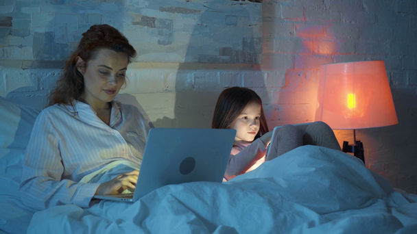 mother using laptop while daughter using digital tablet in bed at night - Imágenes, Vídeo