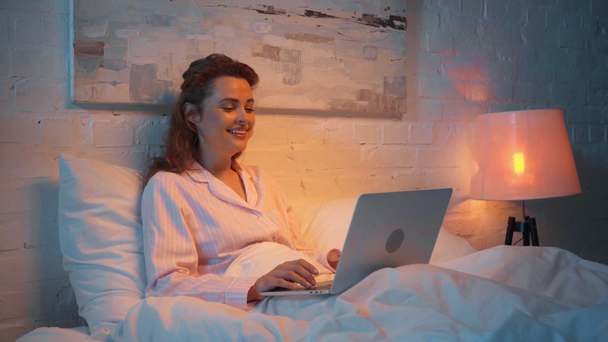 Žena, která se směje při sledování filmu na přenosném počítači v noci v posteli - Záběry, video
