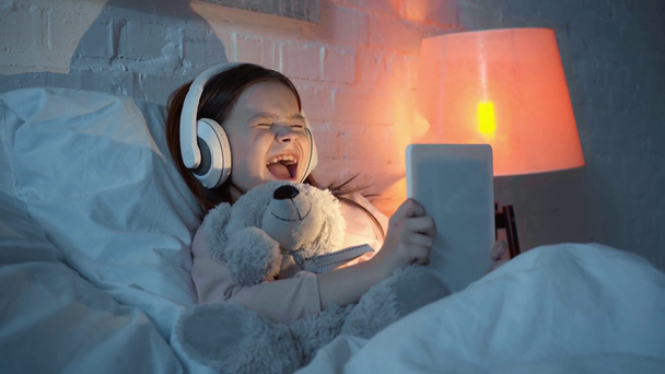 child listening music in headphones, singing and using digital tablet in bed - Footage, Video