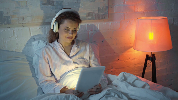 Nainen kuuntelee musiikkia kuulokkeilla ja käyttää digitaalista tablettia sängyssä
 - Materiaali, video