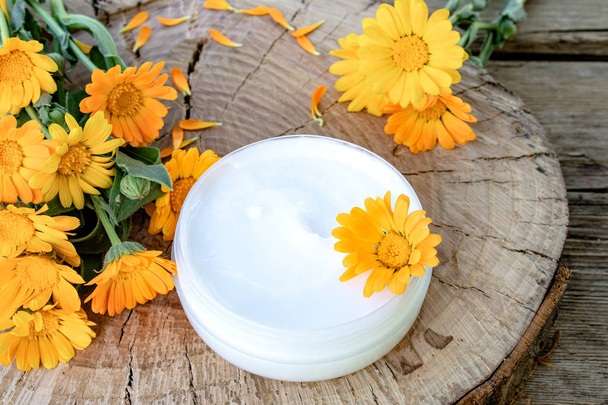 ein Glas weißer Kosmetikcreme für die Körperpflege. frische orange Ringelblumen auf hölzernem Hintergrund. - Foto, Bild