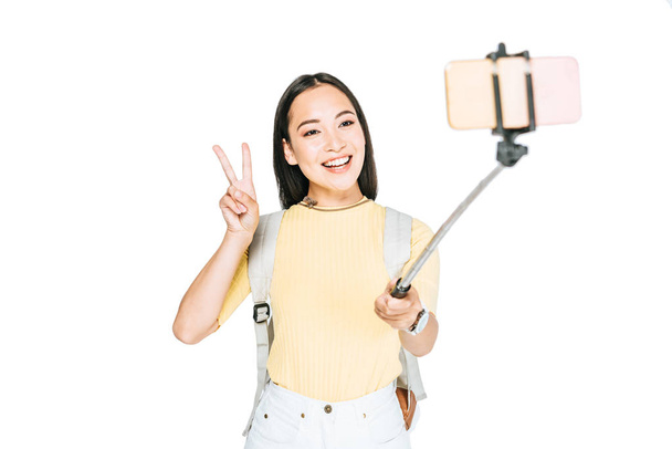 attractive asian woman showing victory sign while taking selfie on smartphone with selfie stick isolated on white - Foto, immagini