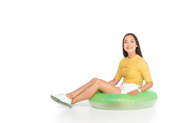 cheerful asian woman sitting on swim ring and smiling at camera on white background - Photo, Image