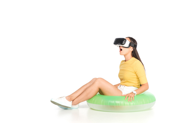 sorprendido asiático mujer usando virtual realidad auricular mientras sentado en nadar anillo en blanco fondo
 - Foto, Imagen