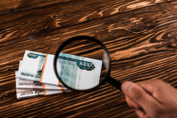cropped view of man holding magnifier near russian money on wooden table  - Photo, Image