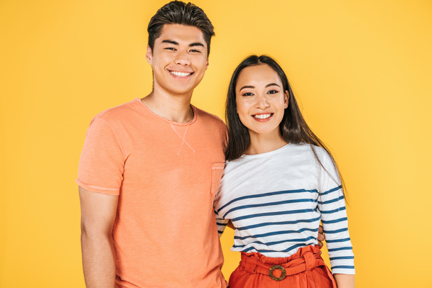 cheerful asian couple embracing while smiling at camera isolated on yellow - Fotografie, Obrázek