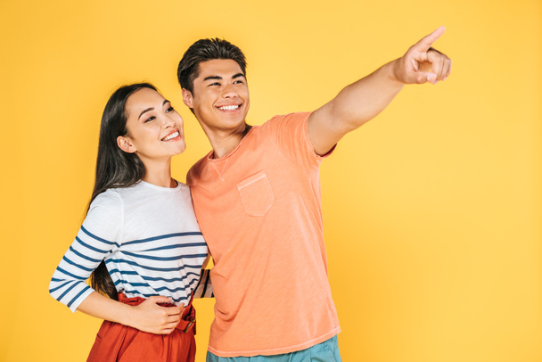 happy asian man looking away and pointing with finger while standing near smiling girlfriend isolated on yellow - Foto, afbeelding