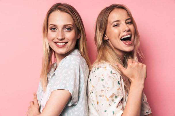 Cheerful woman pointing to her happy pretty blonde women friend sister posing isolated over pink wall background. - Foto, imagen