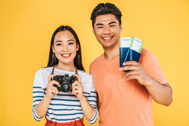 pretty asian girl with digital camera and man with passports and air tickets smiling at camera isolated on yellow - Photo, Image
