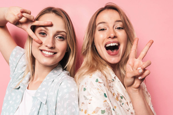 Cheerful pleased beautiful blondes women friends posing isolated over pink wall background showing peace gesture. - 写真・画像
