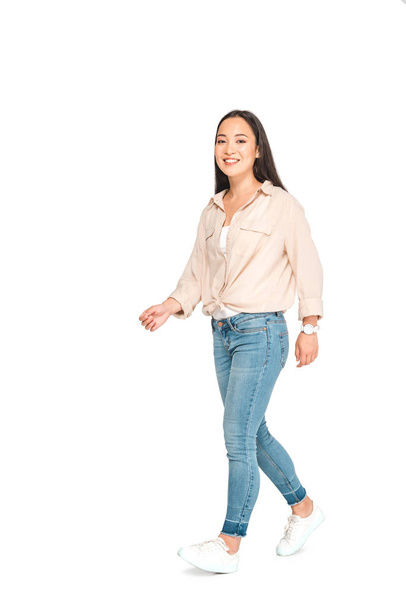 attractive asian woman in blue jeans looking at camera while walking on white background - Фото, зображення