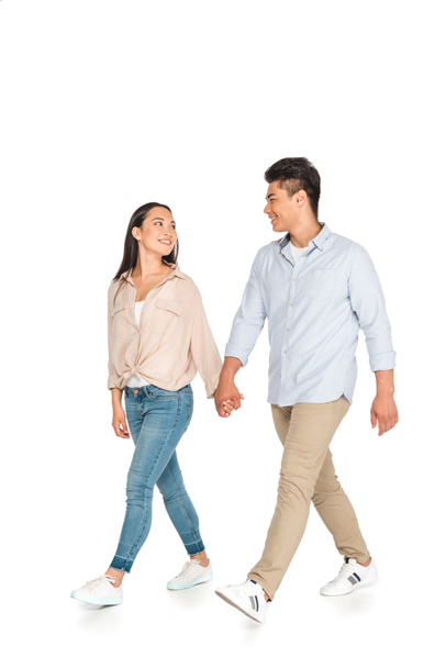 happy asian couple holding hands and looking at each other while walking on white background - Photo, Image