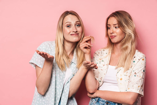 Sad woman holding hair of her happy pretty blonde women friend sister posing isolated over pink wall background. - Zdjęcie, obraz