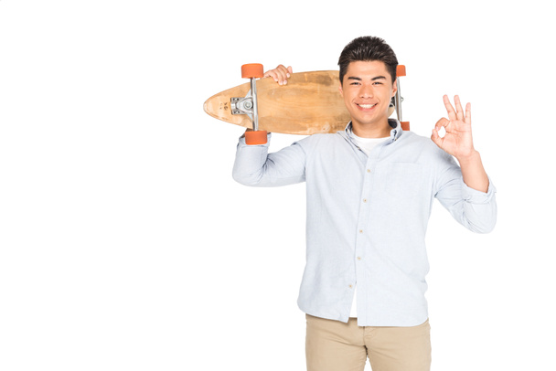 vrolijke Aziatische man houdt Longboard en toont oke teken geïsoleerd op wit - Foto, afbeelding