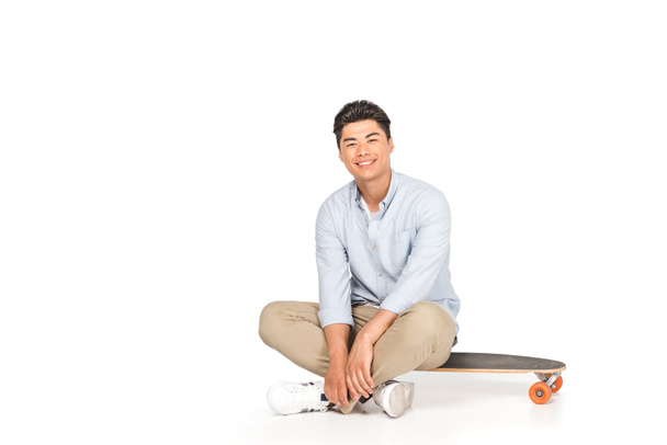 lächelnder asiatischer Mann sitzt auf einem Longboard und blickt in die Kamera auf weißem Hintergrund - Foto, Bild
