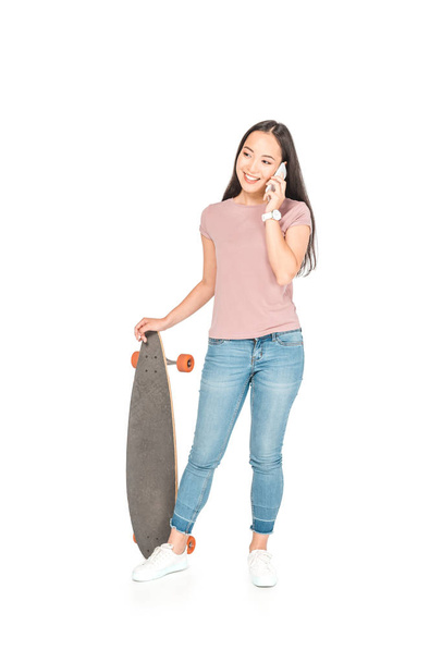 pretty asian girl talking on smartphone while holding longboard on white background - Photo, image